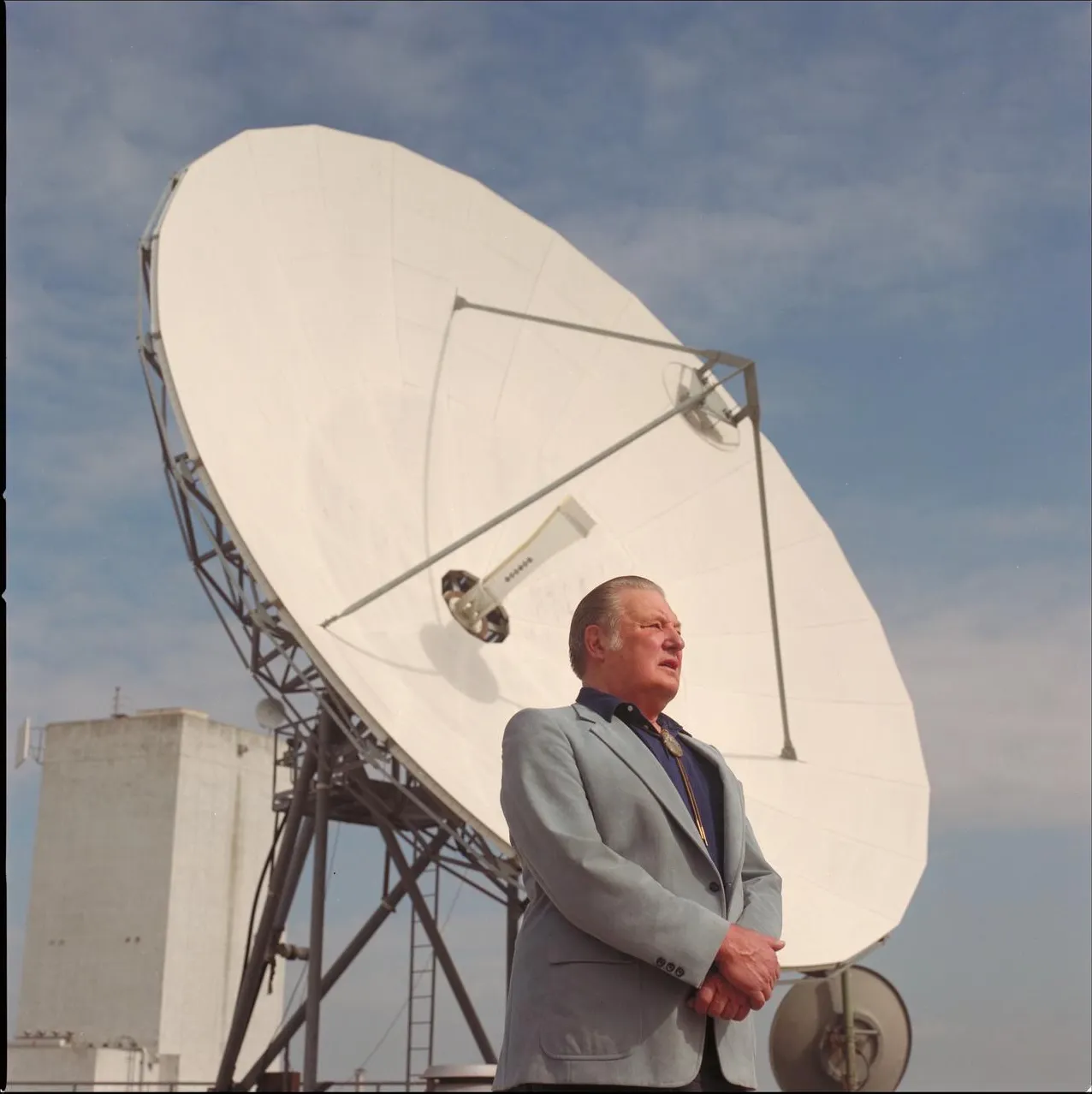 Bernard Oliver in a light jacket standing in front of a white dish antenna under a blue sky