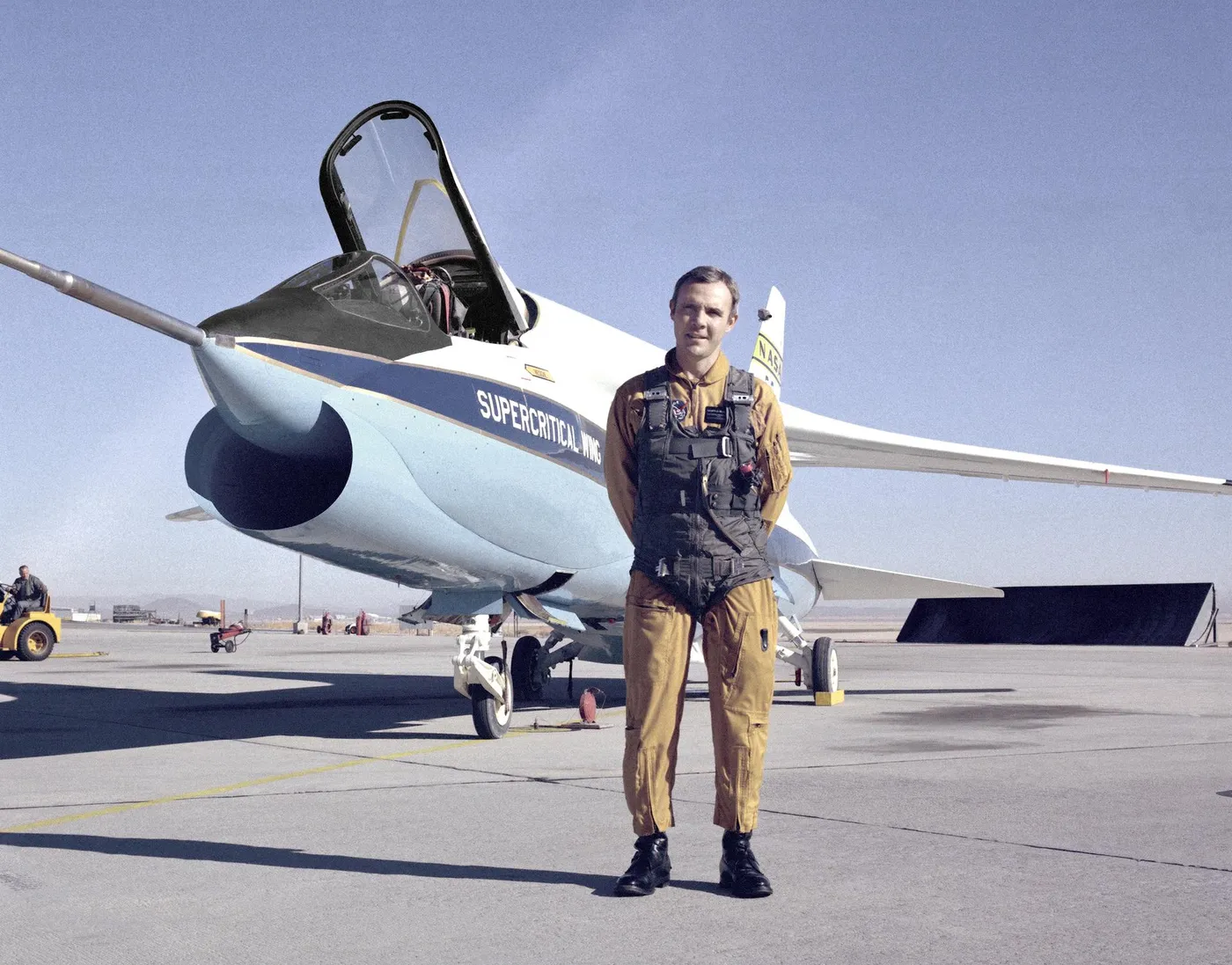 A white and blue F-8 jet with 'Supercritical Wing' on its nose parked on a ramp, with a pilot in a flight suit standing in front