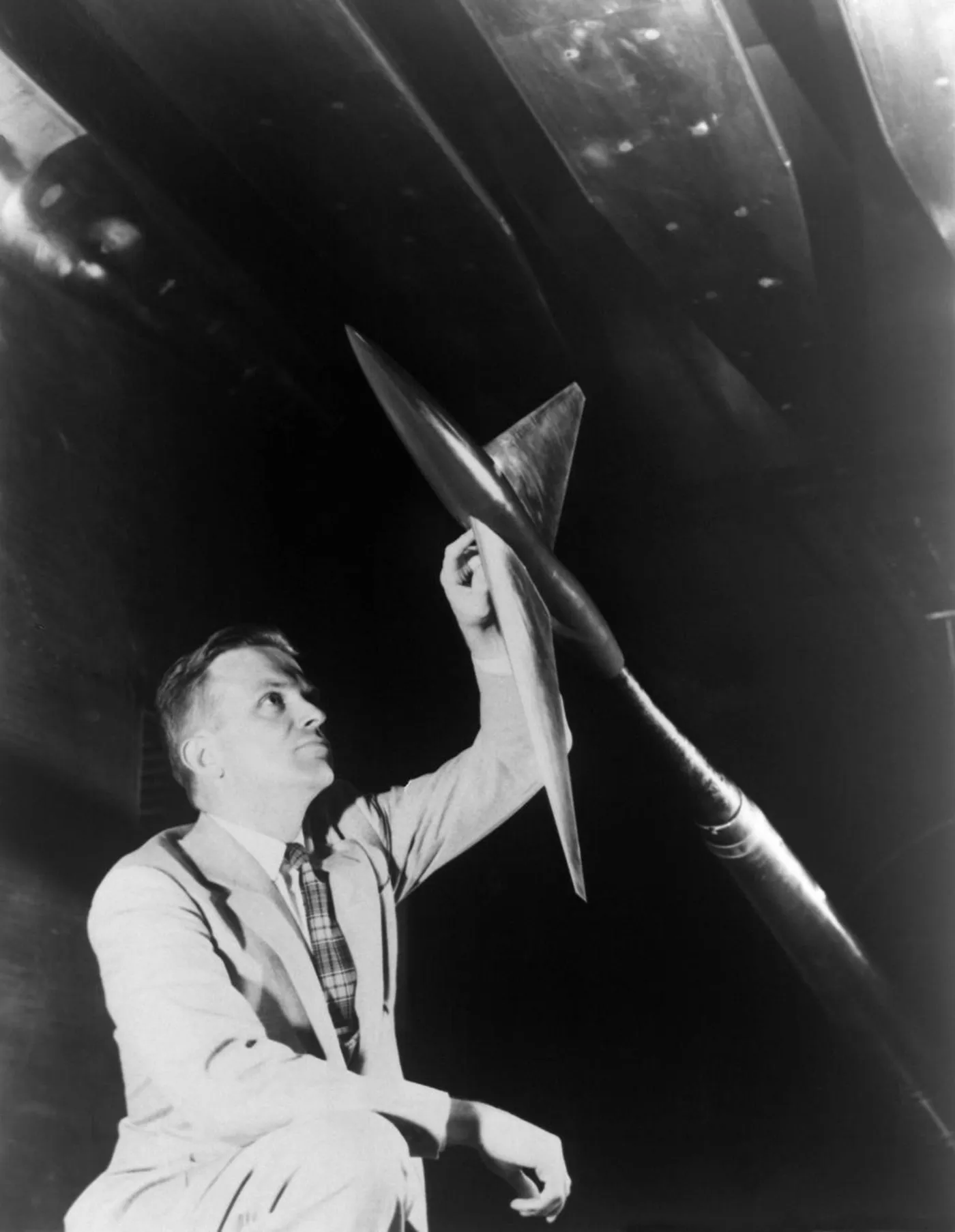 Richard Whitcomb, kneeling in a wind tunnel, reaches up to a sting-mounted model with swept wings and a pinched fuselage
